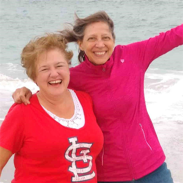 Peggy and Carol have their arms around each other on a beach.