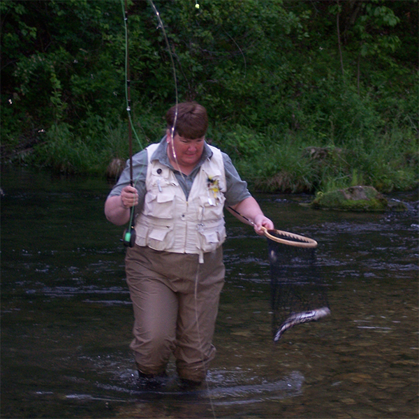 Susan stands in a river holding a fishing rod and a net with a fish in it.